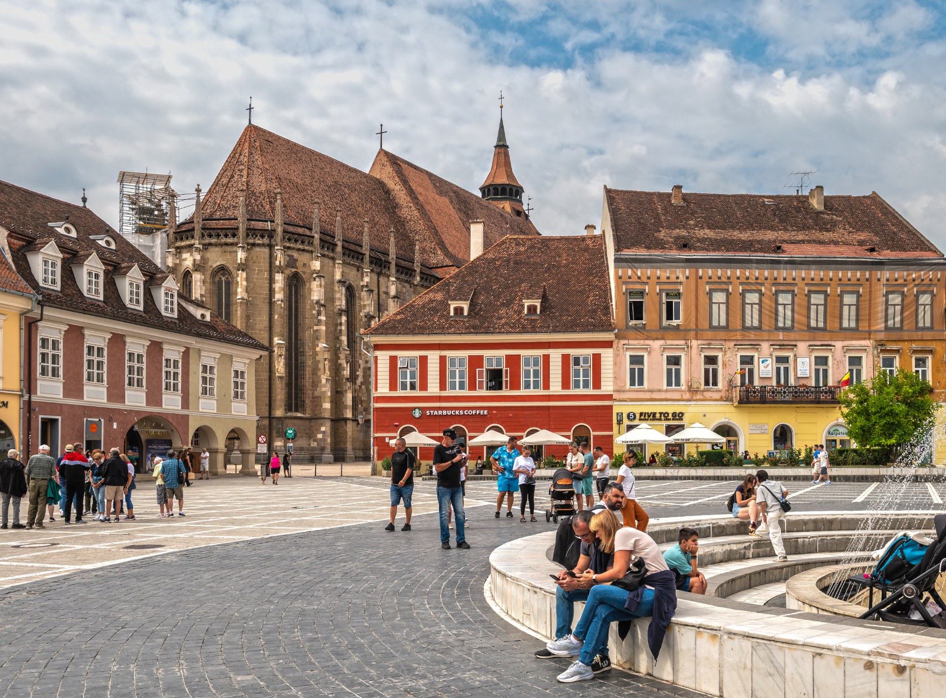 Des jeunes se détendent près de la fontaine sur la place du Conseil à Brașov, avec l’Église Noire en arrière-plan